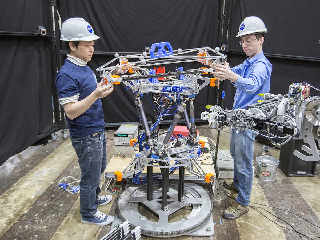 Two NASA engineers work with robotic arms to assemble a metal truss structure.
