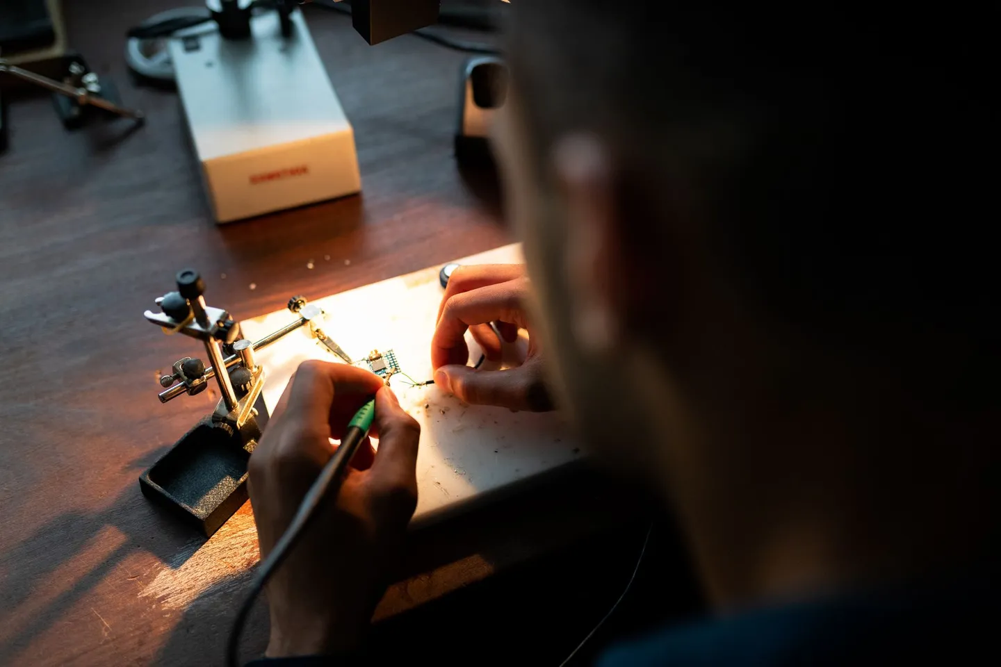 Engineer soldering wires to a small circuit board under a bench lamp, seen over the shoulder